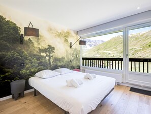 A bedroom with a large bed, a view of a mountainous landscape through a sliding door, and a mural of a forest on the wall.