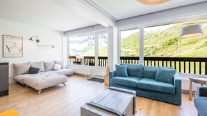 A modern living room with a sofa, a coffee table, and a view of a mountainous landscape through large windows.