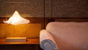 A lamp with a white shade on a wooden nightstand next to a beige armchair.