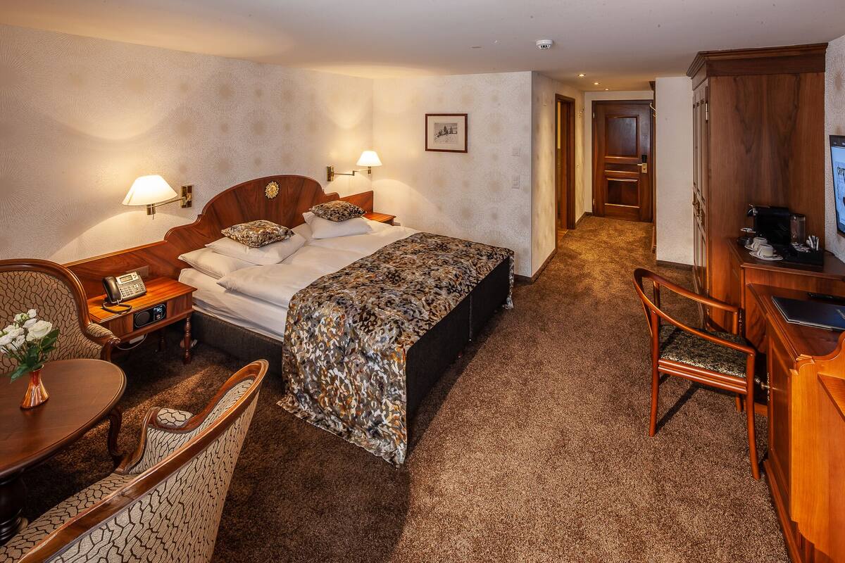 Een hotelkamer met een bed, bedlampjes, een bureau met telefoon, een stoel, een klein tafeltje met vaas en een televisie.