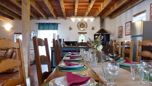 Un espace repas avec une longue table en bois dressée pour le repas, des chaises en bois, une cuisine en arrière-plan et un plafond rustique.