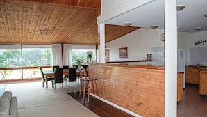 A modern kitchen with wooden cabinetry, a dining area with a table and chairs, and a view through large windows. - Bluebush Estate - The Lodge (Rothbury)