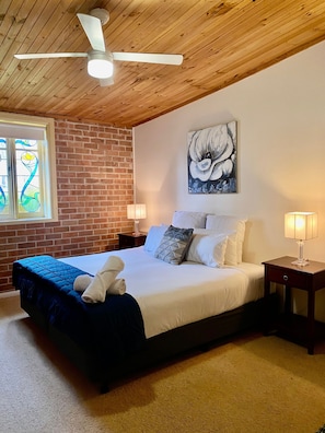 A bedroom with a bed, two bedside tables, a ceiling fan, a stained glass window, and a painting on the wall. - Bluebush Estate - The Lodge (Rothbury)