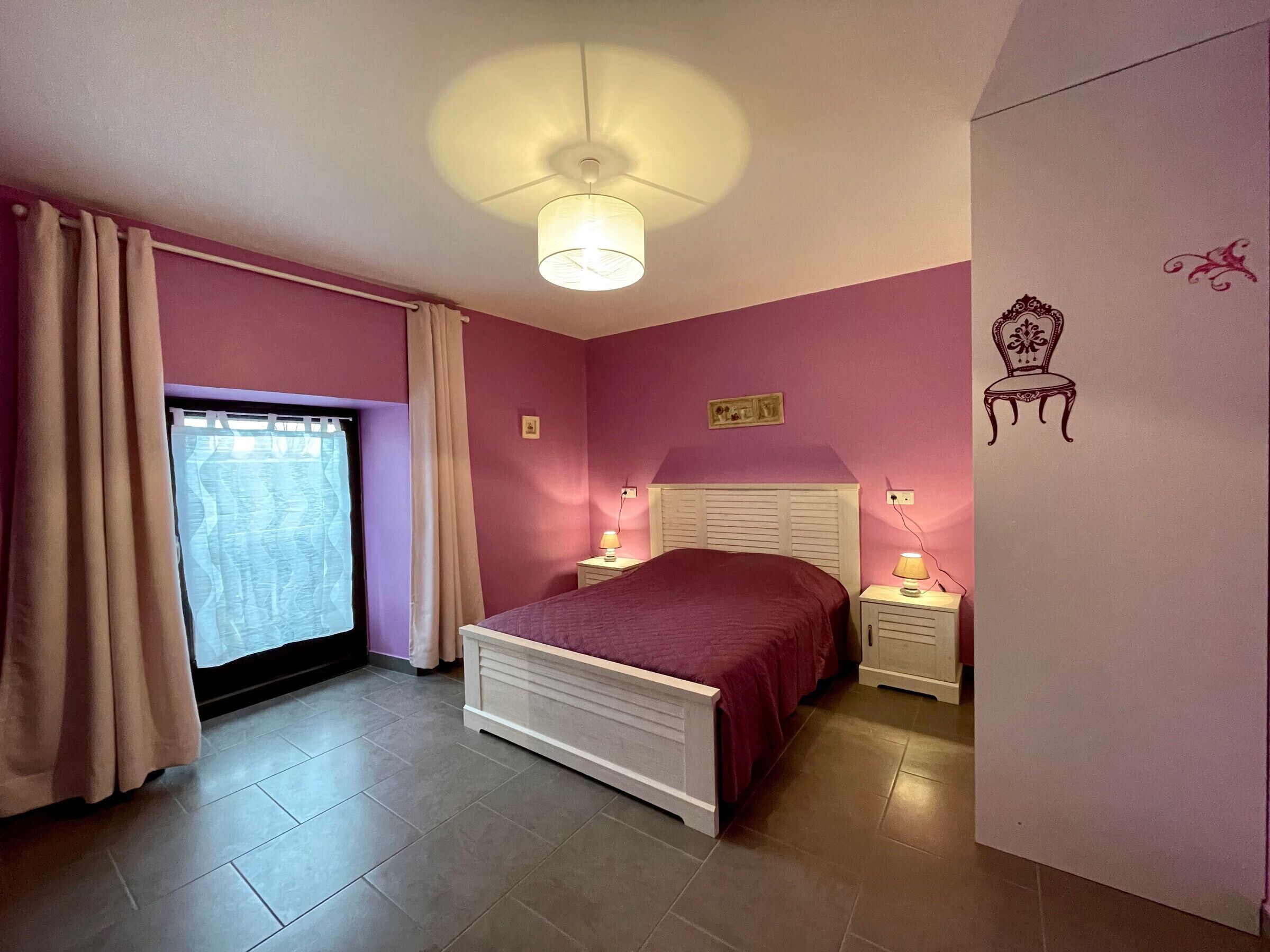 A bedroom with a bed, two bedside tables, a window with curtains, a ceiling light, and a wall decoration.