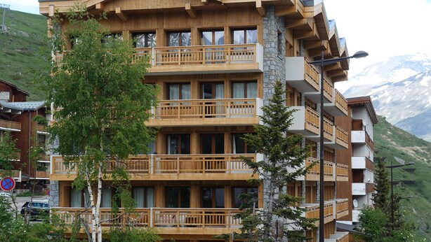 Un hotel de varios pisos con balcones de madera, fachada de piedra y montañas al fondo.
