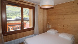 Un dormitorio con paneles de madera, una cama, un ventanal con vistas a un edificio y un paisaje, y una lámpara colgante.