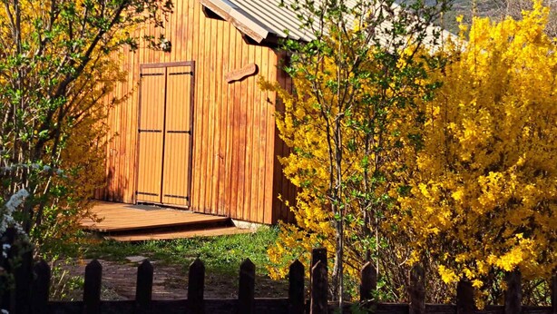 Puinen mökki, jonka katto on tehty metallista, ja jonka ympärillä on puita, joiden lehdet ovat keltaisia. Taustalla näkyy vuoristo.