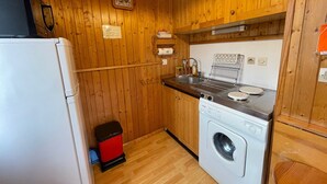 Pieni, käytännöllinen keittiö, jonka seinät ovat puuta, ja jossa on pyykinpesukone, pesuallas ja pieni roskakori.