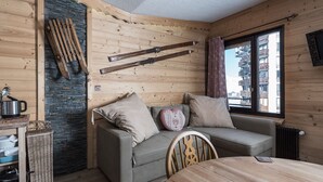 A cozy cabin-style room with a wooden interior, a stone accent wall, a sofa with cushions, a dining table, and a window with a view of buildings.