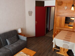 Um espaço de estar compacto com sofá cinzento, uma pequena mesa de madeira, uma mesa de jantar com toalha branca e uma porta vermelha que dá para uma área de cozinha.