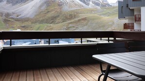 Terraza de madera con una mesa y sillas, con vista a un paisaje montañoso y un edificio con balcones.