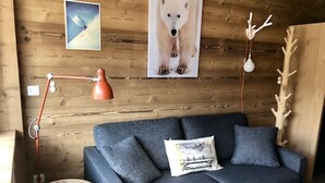 Una habitación acogedora con un sofá azul oscuro, una mesita redonda de madera, una lámpara de pie roja, un póster de un oso polar y una pared de madera con un accesorio de iluminación.