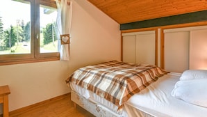 Ein Schlafzimmer mit Holzboden, einem Fenster mit Blick auf Bäume, einem Bett mit karierter Decke und einem Nachttisch.
