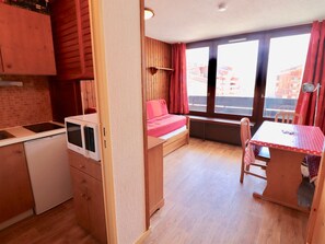 Een kleine kamer met een kitchenette, een bed, een tafel en een raam met gordijnen.