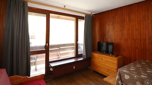 A room with wooden walls, a large window with curtains, a bed, a dresser with a television, and a table with a patterned tablecloth.