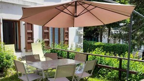 En udendørs terrasse med et bord, stole og en stor parasol, samt udsigt til en bygning med træpersienner.