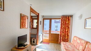 A living room with a sofa, a TV on a cabinet, a small table, and a door leading to a balcony.
