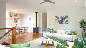Ein geräumiges Wohnzimmer mit weißem Sofa, einem Couchtisch und einem Kamin. Es gibt einen Essbereich mit Holztisch und -stühlen sowie eine Treppe mit Holzwand.