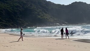 Ein Strand mit Wellen, eine Person geht spazieren und eine andere steht mit einem Surfbrett.