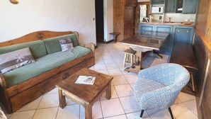 A cozy living room with a sofa, a patterned armchair, a wooden coffee table, and a kitchen area with blue cabinets and a tiled floor.