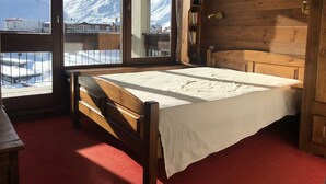 A wooden room with a bed, a window with a view of snow-covered mountains, and a wooden bookshelf.