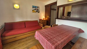 A small, cozy room with a red sofa, a wooden table, and a bed with a checkered bedspread. There is a painting on the wall and a mirror above the bed.