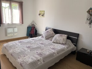 Un dormitorio con cama, mesita de noche, calefactor en la pared, ventana con cortinas y un adorno en la pared.