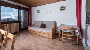 A cozy room with a wooden ceiling, a sofa bed, a small table, and a view of a snowy landscape through a window.
