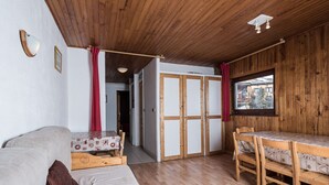 A cozy room with a wooden ceiling, a bed with a patterned bedspread, a dining table with chairs, and a window with a view of buildings.