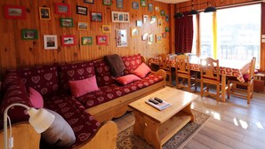 Una sala de estar acogedora con una pared de madera decorada con marcos, un sofá rojo estampado, una mesa de centro de madera y un comedor con sillas y una mesa de madera.
