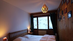 Un dormitorio con cabecera de madera, una cama con ropa de cama blanca, una lámpara de noche, una ventana con vistas a un paisaje nevado y una pared de madera con cuadros.