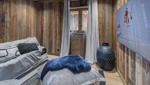 Um quarto aconchegante com paredes de madeira, uma cama com manta azul e branca, um sofá e uma janela grande com cortinas.