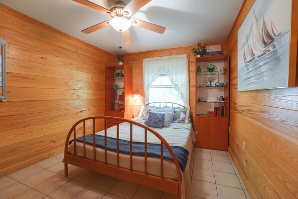 Un dormitorio con cama de madera, ventilador de techo, ventana con cortinas translúcidas y una estantería.