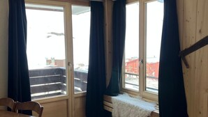 Une pièce dotée d’un plancher en bois, d’une table à manger, de deux chaises, d’un banc recouvert d’un plaid en fourrure et de grandes fenêtres avec des rideaux.