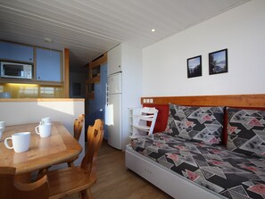 Quarto compacto com mesa de jantar de madeira, cadeiras, sofá com almofadas estampadas, micro-ondas e frigorífico.