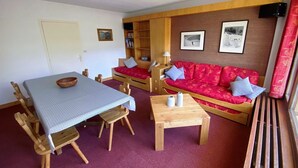A cozy living room with a dining table, wooden chairs, a red sofa with cushions, a bookshelf, and framed pictures on the wall. - Beautiful apartment for 7, balcony, direct slope access (Tignes)