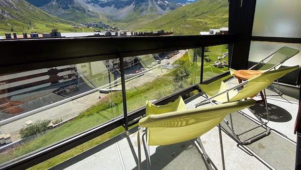 A balcony with a yellow chair, a glass railing, and a view of a mountainous landscape with a road and buildings. - Beautiful apartment for 7, balcony, direct slope access (Tignes)