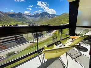 Un balcón con una silla amarilla, barandilla de vidrio y vista a un paisaje montañoso con carretera y edificios.