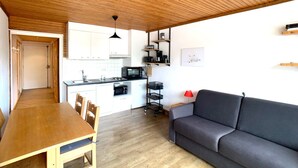 A compact living space with a wooden ceiling, a kitchenette, a dining area with a table and chairs, and a grey sofa.