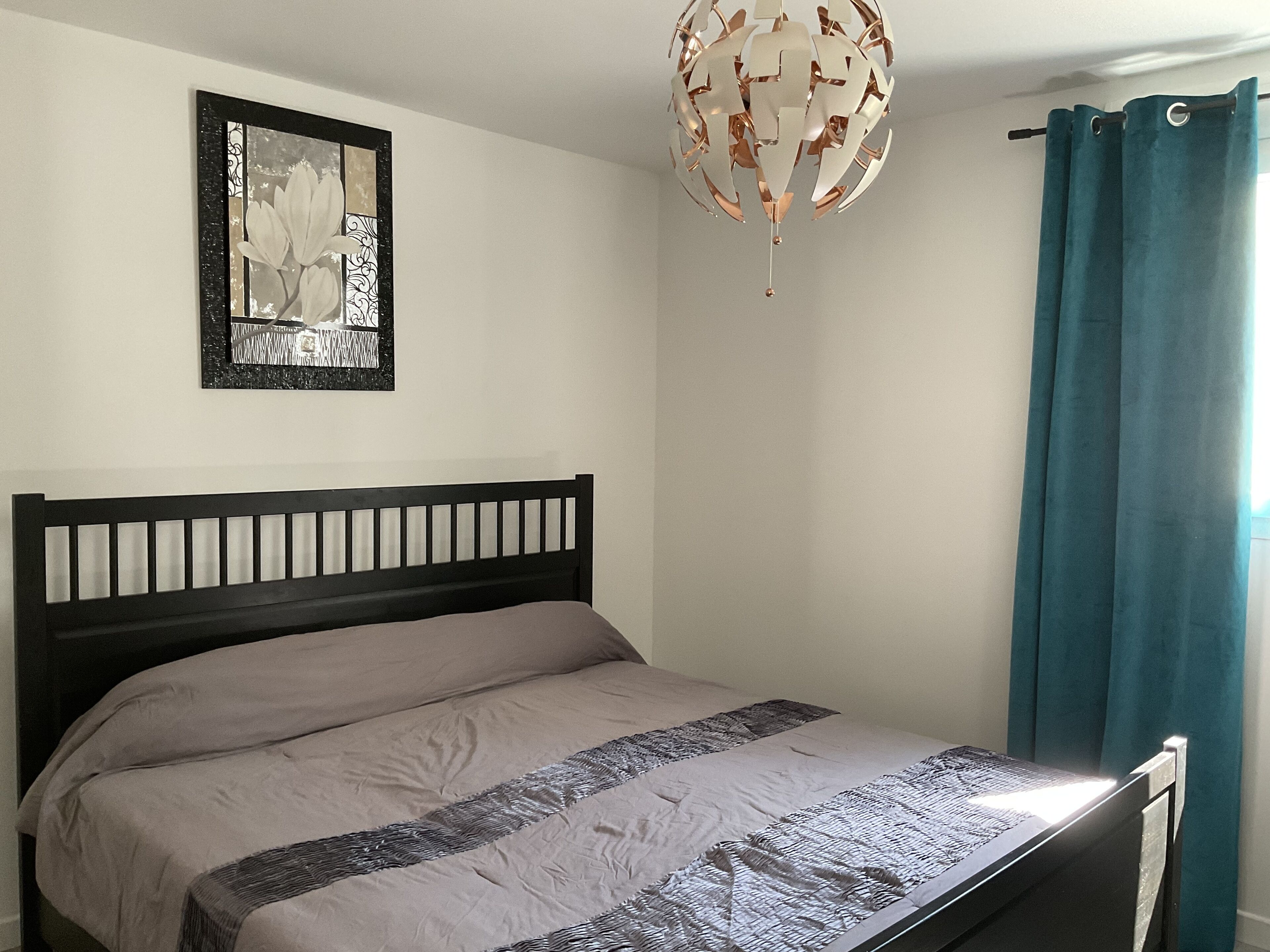 Una camera da letto con un letto, una lampada a sospensione, un quadro appeso al muro e una finestra con tende.