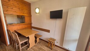 A small, cozy room with a wooden table and chairs, a small stool, a television mounted on the wall, and a cabinet with a mirror.