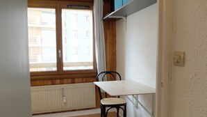 Um quarto pequeno com janela, um radiador, uma cadeira, uma mesa e um banquinho azul.