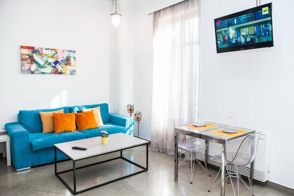 A modern living room with a blue sofa, a coffee table, and a dining table with chairs. A flat-screen TV is mounted on the wall. - Beautiful Apt. close to Monastiraki (Athens)