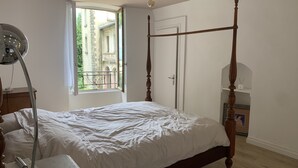 Ein Bett mit Baldachin, eine weiße Bettdecke, eine Nachttischlampe und Blick auf einen Balkon mit Grünpflanzen.