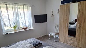 Ein Schlafzimmer mit zwei Betten, einem Sessel, einem Kleiderschrank, einem Fernseher und einem Fenster mit leichtem Vorhang.
