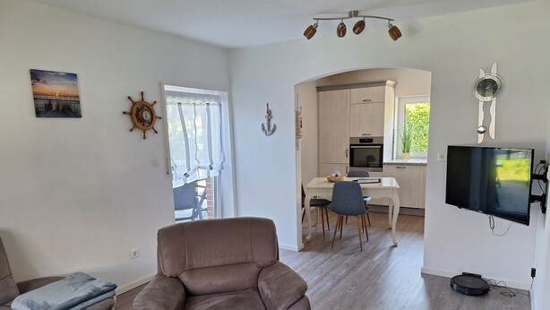 Ein modernes Wohnzimmer mit einer Couch, einem Essbereich mit Tisch und Stühlen, einer Küche mit Geräten und einem an der Wand montierten Fernseher.