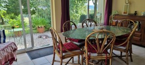 A dining area with a glass table surrounded by wicker chairs, a view of a garden through large windows, and a wooden cabinet with decorative items. - Pech Ferrat, Pleine Campagne sur 1 Ha Arboré, vue sur la Vallée et les Coteaux (Saint-Amans-du-Pech)