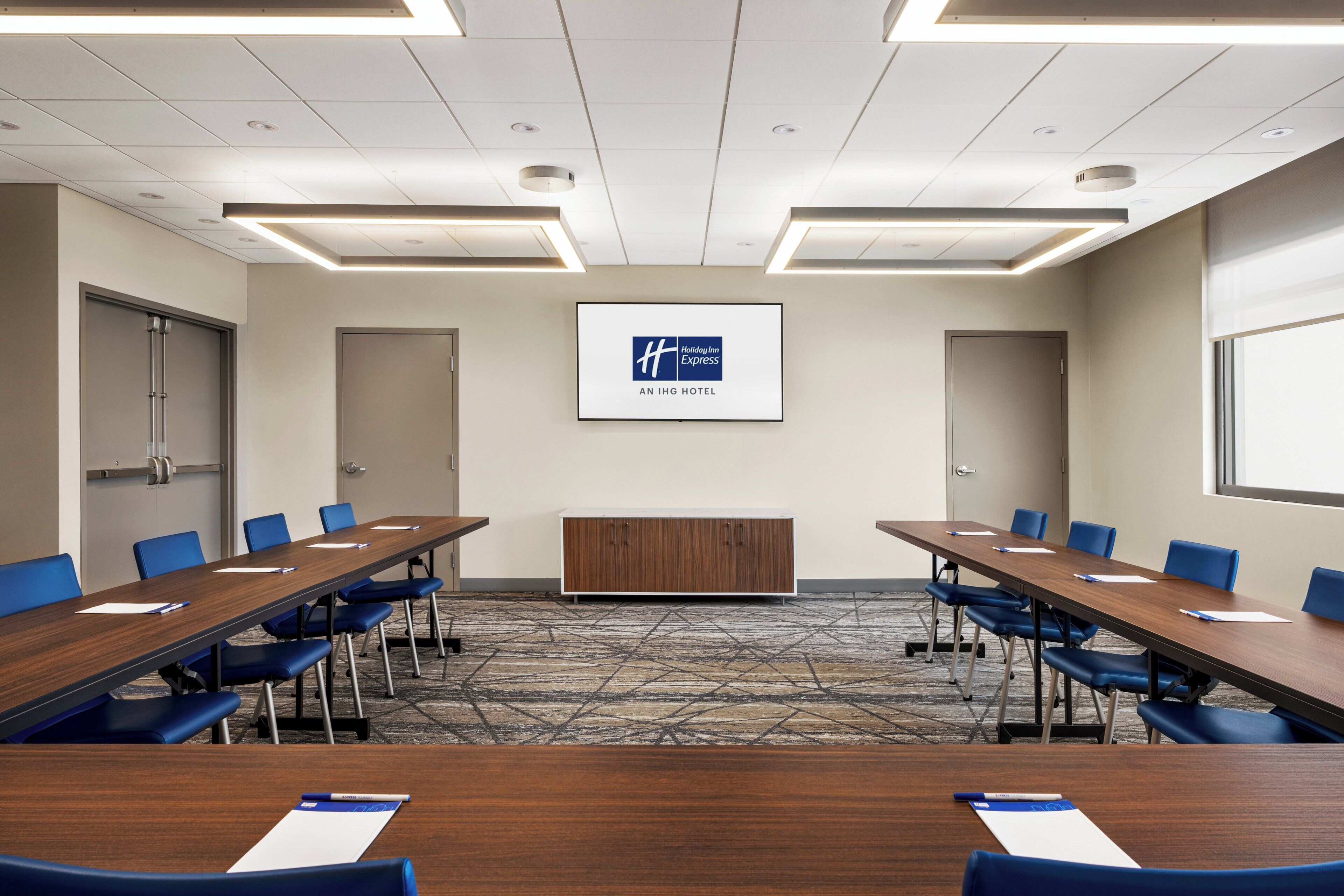 A conference room with a long table, blue chairs, a podium, and a screen displaying the hotel name.