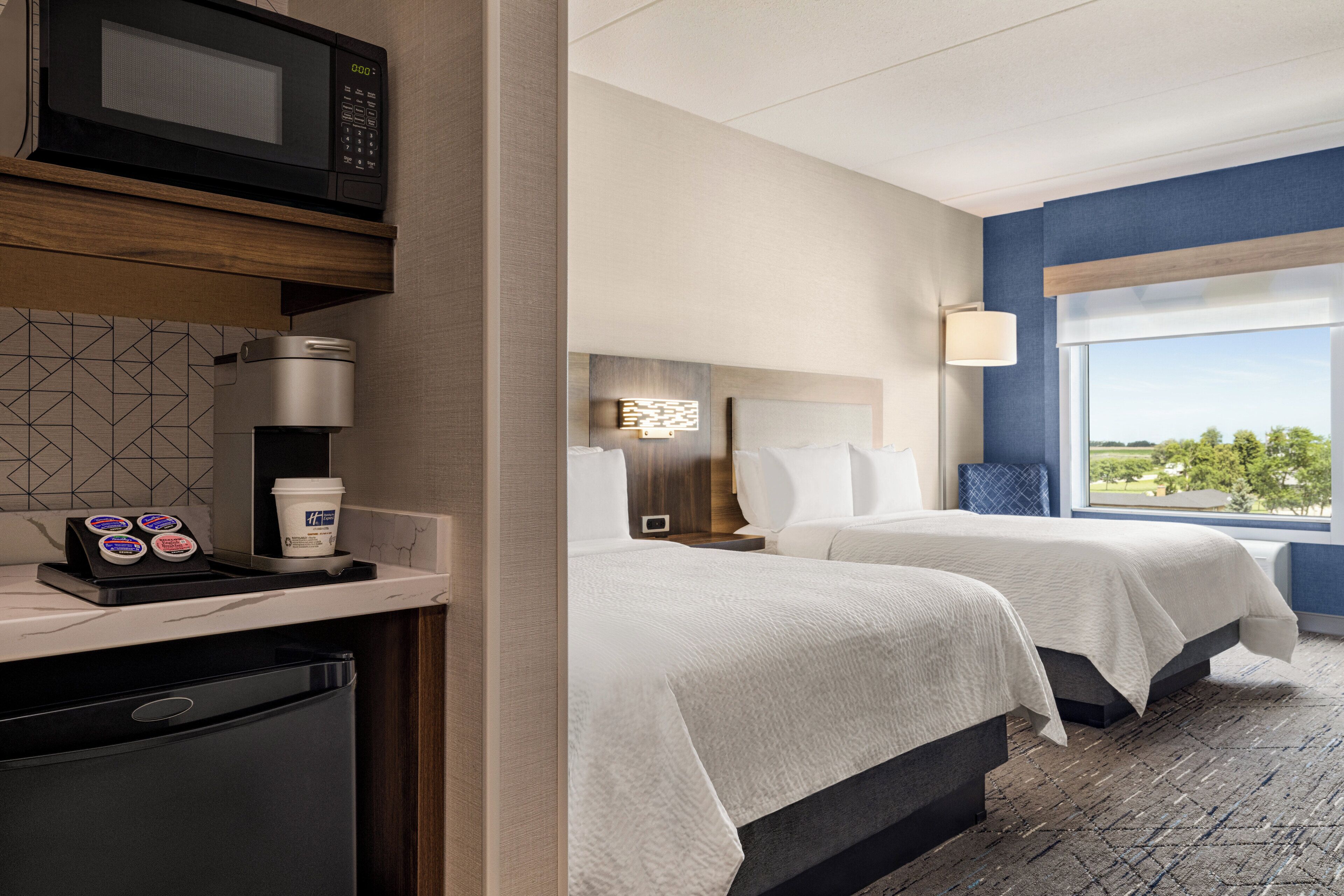 A hotel room with a microwave, coffee maker, and a small refrigerator. There are two beds with white linens and a large window offering a view of the outdoors.