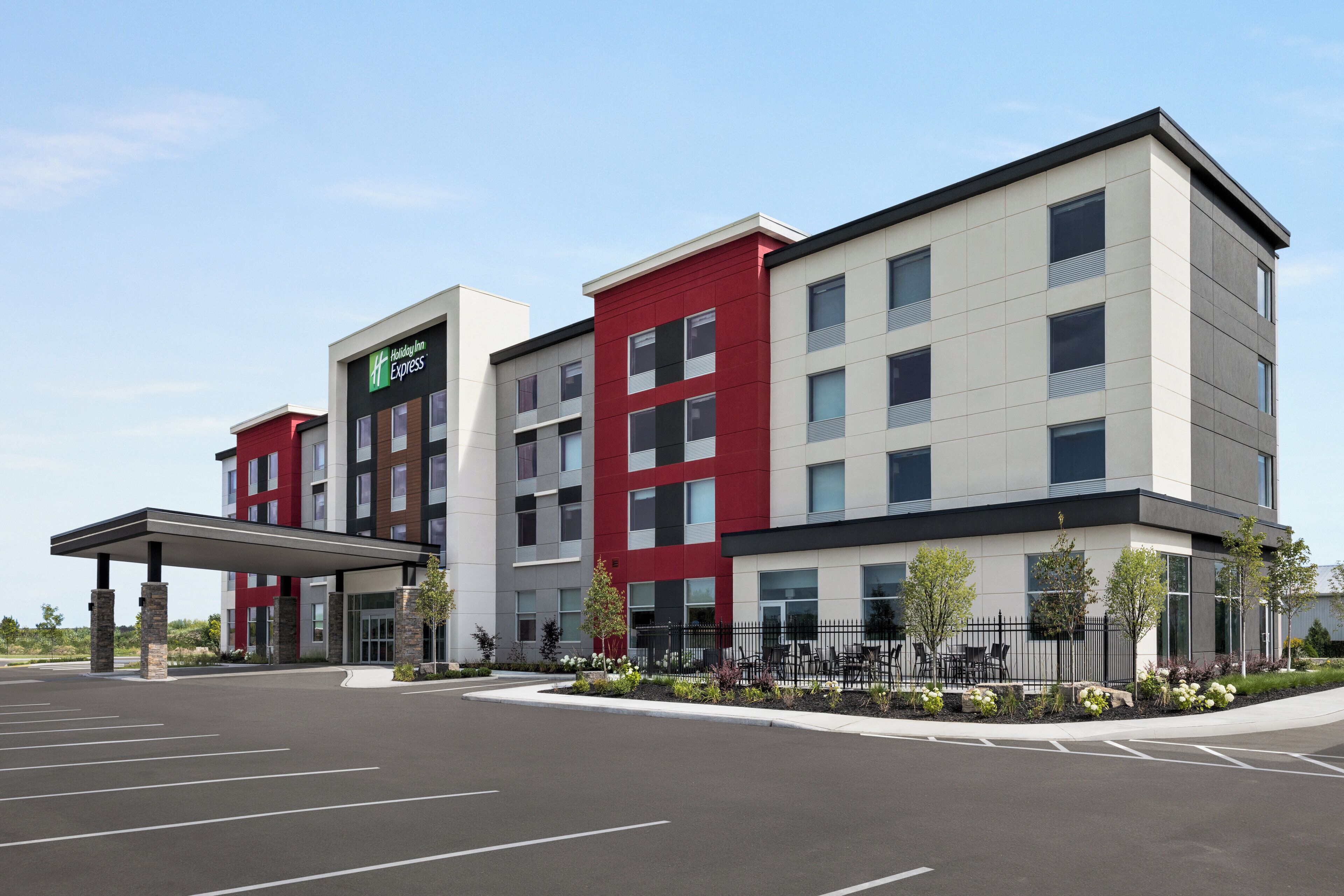 A multi-story hotel with a modern design, featuring a mix of red and neutral colors, a covered entrance, and a landscaped outdoor area with a fence and seating.